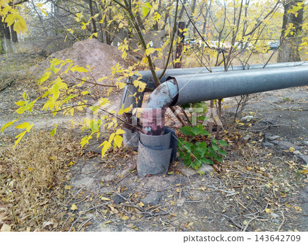 Weathered metal utility pipes with damaged insulation elbows run along the ground in a neglected, autumnal outdoor setting near trees. Weathered metal utility pipes with damaged insulation elbows run along the ground in a neglected, autumnal outdoor setting near trees. 143642709