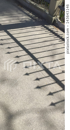 Sharp shadows of a metal fence with pointed finials stretch across an asphalt sidewalk. Sharp shadows of a metal fence with pointed finials stretch across an asphalt sidewalk. 143643146