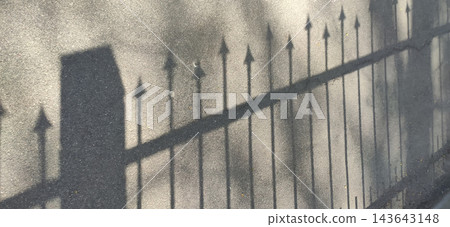 Sharp shadows of a metal fence with pointed finials stretch across an asphalt sidewalk. Sharp shadows of a metal fence with pointed finials stretch across an asphalt sidewalk. 143643148