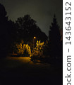 An autumn park at dusk with illuminated lanterns, colorful trees, and fallen leaves covering the paths. 143643152