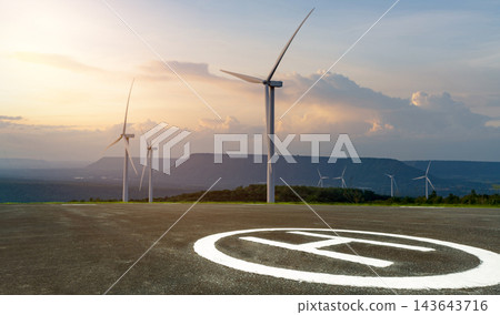 Asphalt helipad overlooking wind turbine farm at sunset, renewable wind energy infrastructure generating clean electricity beside helicopter landing platform for sustainable aviation and transport Asphalt helipad overlooking wind turbine farm at sunset, renewable wind energy infrastructure generating clean electricity beside helicopter landing platform for sustainable aviation and transport 143643716