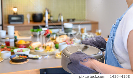 一位身穿圍裙的女士在廚房裡端著一個鍋，桌上擺放著派對食物。 143644198