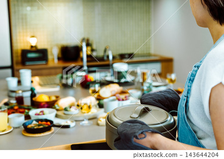 一位身穿圍裙的女子在廚房裡端著鍋,餐桌上擺著食物。 一位身穿圍裙的女子在廚房裡端著鍋,餐桌上擺著食物。 143644204