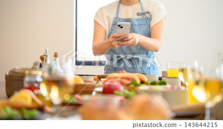 一位身穿圍裙的女士正在用智慧型手機拍攝她做飯的照片。 一位身穿圍裙的女士正在用智慧型手機拍攝她做飯的照片。 143644215