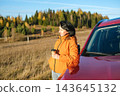 Woman leaning on red car holding coffee paper cup during autumn road trip, resting and looking at scenic landscape. Travel and freedom concept 143645132