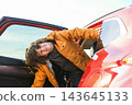 Playful boy leaning out of open red car door during family road trip, cheerful child traveler in jacket outdoors, vacation and journey concept 143645133