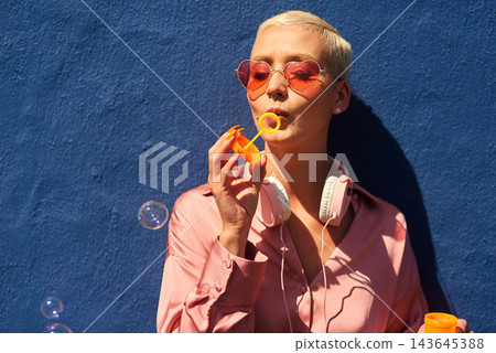 Young, woman and blowing bubbles against wall for fashion or wellness, cool with heart sunglasses for trend. Female person, outdoor and backdrop with sunshine, headphones and clothes for style in USA Young, woman and blowing bubbles against wall for fashion or wellness, cool with heart sunglasses for trend. Female person, outdoor and backdrop with sunshine, headphones and clothes for style in USA 143645388