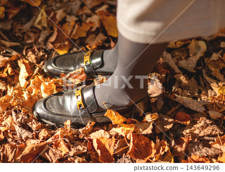 Black loafers with gold chain buckle, woman walking on autumn leaves. Stylish footwear detail, gray tights, fallen foliage on ground. Fashion close up, trendy metallic accessory, fall mood. Black loafers with gold chain buckle, woman walking on autumn leaves. Stylish footwear detail, gray tights, fallen foliage on ground. Fashion close up, trendy metallic accessory, fall mood. 143649296