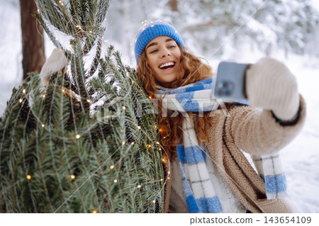 A young woman with a phone and a Christmas tree with garlands enjoys the beautiful winter weather. A young woman with a phone and a Christmas tree with garlands enjoys the beautiful winter weather. 143654109