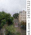 Abstract view of raindrops on a glass pane with an overcast, moody bokeh of green trees and urban buildings in the background. 143657122