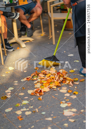 Sweeping Autumn Leaves on Outdoor Pavement. Autumn leaves are swept into pile with broom on paved outdoor cafe terrace, seasonal cleanup. Fallen leaves cover pavement around seating area Sweeping Autumn Leaves on Outdoor Pavement. Autumn leaves are swept into pile with broom on paved outdoor cafe terrace, seasonal cleanup. Fallen leaves cover pavement around seating area 143657536