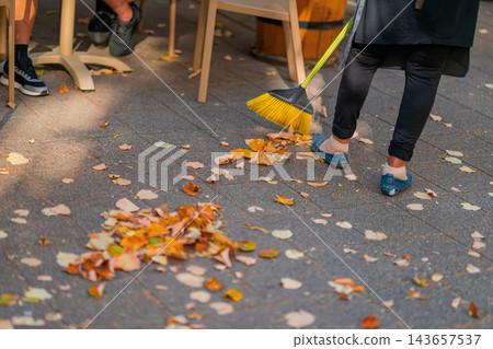 Cleaner sweeping golden autumn leaves with yellow broom on sidewalk. Low angle cropped view of person sweeping fallen yellow autumn leaves off paved city sidewalk near outdoor cafe using bright broom Cleaner sweeping golden autumn leaves with yellow broom on sidewalk. Low angle cropped view of person sweeping fallen yellow autumn leaves off paved city sidewalk near outdoor cafe using bright broom 143657537