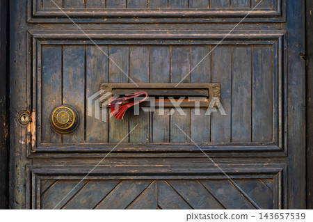 Old Wooden Door With Mail Slot. Vintage door features brass mail slot holding folded printed paper, decorative round hardware. Weathered blue wooden door shows scratched paint. Old Wooden Door With Mail Slot. Vintage door features brass mail slot holding folded printed paper, decorative round hardware. Weathered blue wooden door shows scratched paint. 143657539