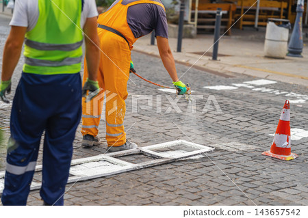 Road workers painting pedestrian crossing lines. Low angle view of construction workers applying white road marking paint with spray gun on cobblestone pavement using template frame on city street Road workers painting pedestrian crossing lines. Low angle view of construction workers applying white road marking paint with spray gun on cobblestone pavement using template frame on city street 143657542
