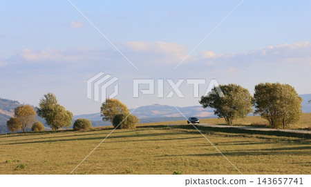 A car travels along a quiet country road through rolling golden hills dotted with autumn trees 143657741