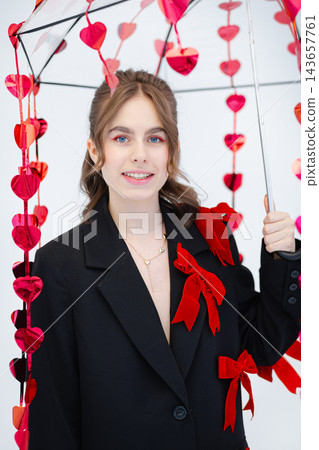 Young woman holding umbrella decorated with red heart garlands wearing black blazer with red bows and heart necklace 143657761