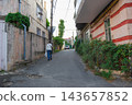 Woman walking up a narrow asphalt street between old residential buildings. 143657852