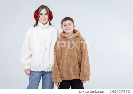 Smiling young girl wearing white fleece jacket and red earmuffs standing next to boy with brown hooded fleece jacket Smiling young girl wearing white fleece jacket and red earmuffs standing next to boy with brown hooded fleece jacket 143657970