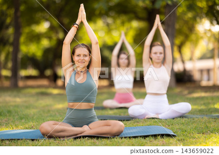 Portrait of women sitting on gym mat and doing yoga meditation in lotus position and arms raised in park Portrait of women sitting on gym mat and doing yoga meditation in lotus position and arms raised in park 143659022