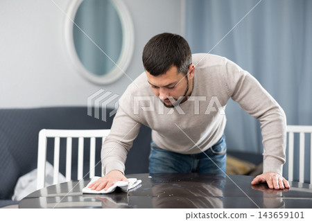 Owner of apartment, man carefully wipes the table in the room at home with rag 143659101
