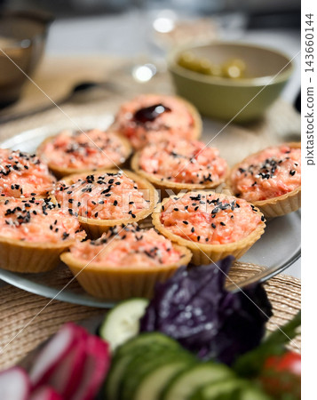 Capelin caviar tartlets topped with black and white sesame seeds sit on plate on kitchen counter. Bowl and sliced vegetables, tasty food and serving, decorative for design. Simple dinner 143660144