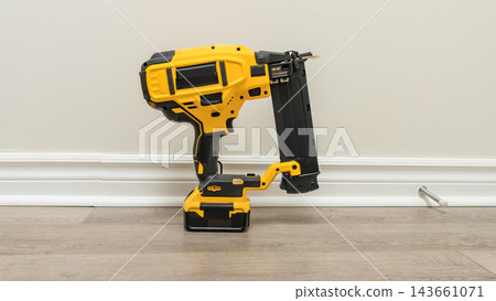 Yellow cordless fastening tool standing upright on a wood floor against a plain wall in an indoor home setting Yellow cordless fastening tool standing upright on a wood floor against a plain wall in an indoor home setting 143661071