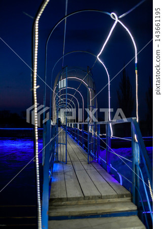 Illuminated pedestrian bridge with a wooden walkway and metal frame, showcasing blue lighting at night over a body of water 143661195