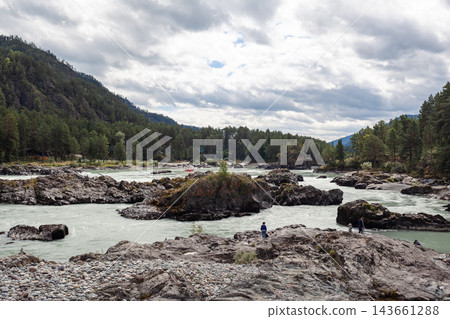 Scenic view of a river with rocky banks, surrounded by dense green forests and mountains under a cloudy sky, featuring people along the shore 143661288