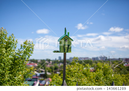 Colorful birdhouse mounted on a post amidst lush green trees, with a clear blue sky and distant buildings visible in the background 143661289