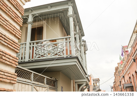 Ornate gray balcony with patterned railing and hanging lights projects between historic brick facades, overlooking a narrow tbilisi street Ornate gray balcony with patterned railing and hanging lights projects between historic brick facades, overlooking a narrow tbilisi street 143661338