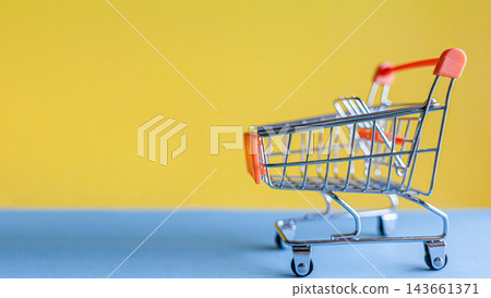 Miniature shopping cart positioned on a blue surface against a bright yellow background, suitable for retail and e-commerce imagery Miniature shopping cart positioned on a blue surface against a bright yellow background, suitable for retail and e-commerce imagery 143661371
