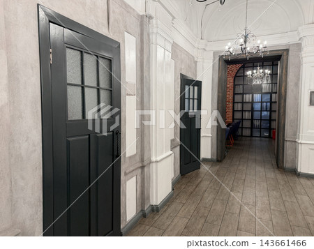 Interior hallway features gray walls and wooden flooring, with black doors on either side leading to rooms and elegant chandeliers above Interior hallway features gray walls and wooden flooring, with black doors on either side leading to rooms and elegant chandeliers above 143661466