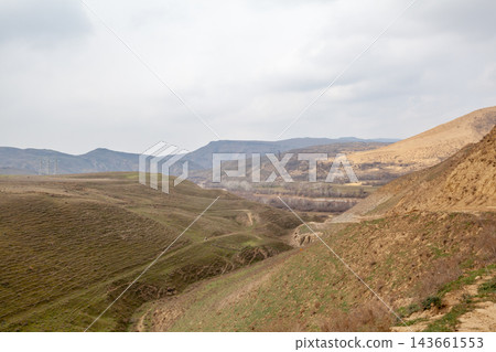 Rolling hills and valleys are visible under a cloudy sky, with distant mountains and sparse vegetation creating a serene rural landscape Rolling hills and valleys are visible under a cloudy sky, with distant mountains and sparse vegetation creating a serene rural landscape 143661553