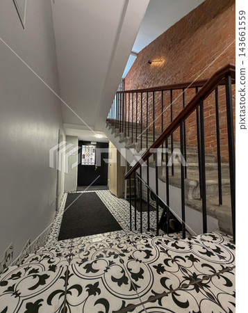 Interior view of a staircase featuring decorative tile flooring and a wooden railing, with an exposed brick wall and a black door at the top Interior view of a staircase featuring decorative tile flooring and a wooden railing, with an exposed brick wall and a black door at the top 143661559