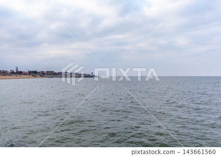 Coastal view showcasing a tranquil sea and a distant shoreline with buildings, set against a backdrop of overcast clouds 143661560