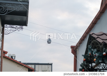 A small cable car cabin travels above red rooflines, decorative wrought iron balconies, and a christmas tree in central tbilisi A small cable car cabin travels above red rooflines, decorative wrought iron balconies, and a christmas tree in central tbilisi 143661704