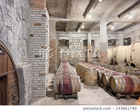 Wooden wine barrels rest on supports throughout a stone and brick cellar, surrounded by concrete columns, textured walls, and industrial lighting 143661740