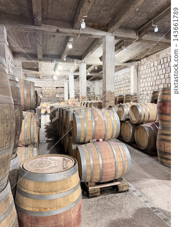 Wooden barrels arranged in rows across an aged stone cellar, with metal bands, concrete beams, exposed masonry walls, and overhead lights Wooden barrels arranged in rows across an aged stone cellar, with metal bands, concrete beams, exposed masonry walls, and overhead lights 143661789