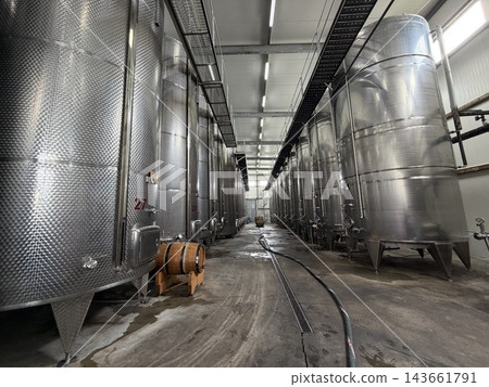 Large stainless steel tanks line both sides of an industrial cellar, with wooden barrels, hoses, overhead walkways, and concrete floors Large stainless steel tanks line both sides of an industrial cellar, with wooden barrels, hoses, overhead walkways, and concrete floors 143661791