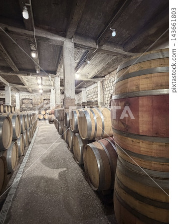 Wooden barrels stand in orderly rows along a narrow stone and concrete cellar, with overhead bulbs illuminating the textured storage interior Wooden barrels stand in orderly rows along a narrow stone and concrete cellar, with overhead bulbs illuminating the textured storage interior 143661803