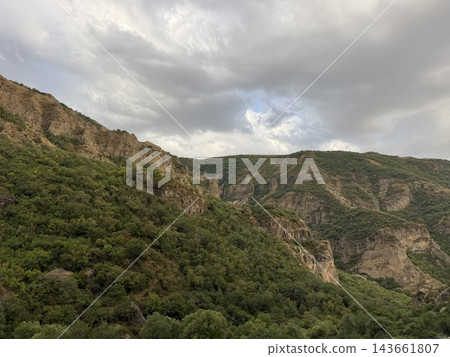Green forested mountains and rugged cliffs fill a broad valley beneath dramatic gray clouds, showing layered terrain and a natural wilderness landscape Green forested mountains and rugged cliffs fill a broad valley beneath dramatic gray clouds, showing layered terrain and a natural wilderness landscape 143661807