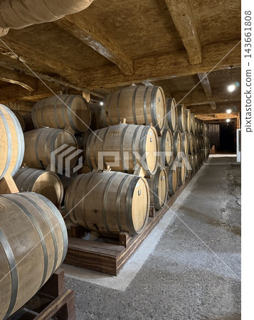 Wooden barrels rest in organized rows beneath rough timber beams, with concrete flooring and warm lights extending through a rustic cellar Wooden barrels rest in organized rows beneath rough timber beams, with concrete flooring and warm lights extending through a rustic cellar 143661808