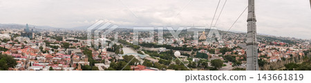 Wide panorama of tbilisi with red roofed buildings, a winding river, wooded areas, rolling hills, and a tall utility pole in view Wide panorama of tbilisi with red roofed buildings, a winding river, wooded areas, rolling hills, and a tall utility pole in view 143661819