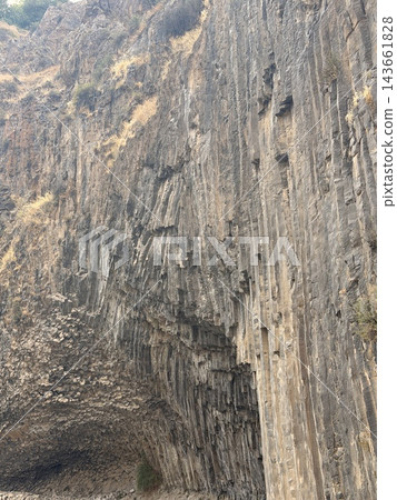 Garni, Armenia - August 26, 2026: Tall basalt columns create a rugged cliff, with weathered gray rock, scattered vegetation, loose stones, and a dark recess Garni, Armenia - August 26, 2026: Tall basalt columns create a rugged cliff, with weathered gray rock, scattered vegetation, loose stones, and a dark recess 143661828
