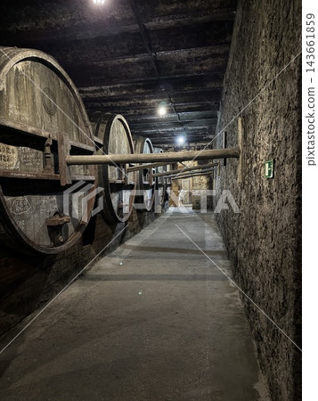 Massive wooden barrels stand along both sides of a narrow stone corridor beneath dark timber beams in an illuminated underground cellar Massive wooden barrels stand along both sides of a narrow stone corridor beneath dark timber beams in an illuminated underground cellar 143661859