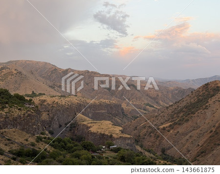 Rugged mountain valley displays layered dry slopes and distant ridges, with scattered green trees and pastel clouds beneath softly lit sky Rugged mountain valley displays layered dry slopes and distant ridges, with scattered green trees and pastel clouds beneath softly lit sky 143661875
