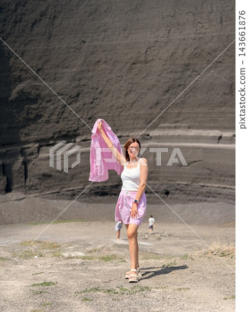 Woman in a white top and pink shorts holds matching patterned fabric before a layered dark rock wall and open gravel ground Woman in a white top and pink shorts holds matching patterned fabric before a layered dark rock wall and open gravel ground 143661876