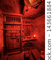 Red illumination reveals stone walls, carved wooden doors, balcony railings, ceiling beams, a small lamp, and rustic furnishings inside a chamber 143661884