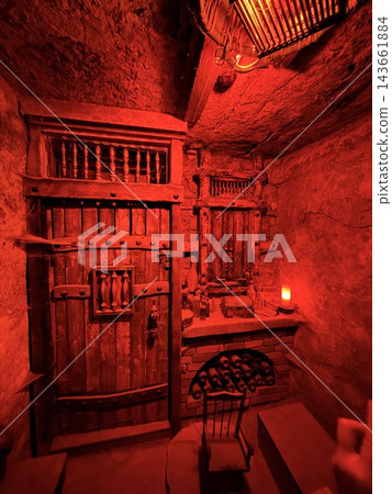 Red illumination reveals stone walls, carved wooden doors, balcony railings, ceiling beams, a small lamp, and rustic furnishings inside a chamber Red illumination reveals stone walls, carved wooden doors, balcony railings, ceiling beams, a small lamp, and rustic furnishings inside a chamber 143661884
