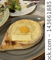 Oval baked bread holds melted cheese, sunny egg yolk, and butter on a ceramic plate, with salad remnants and cutlery nearby 143661885